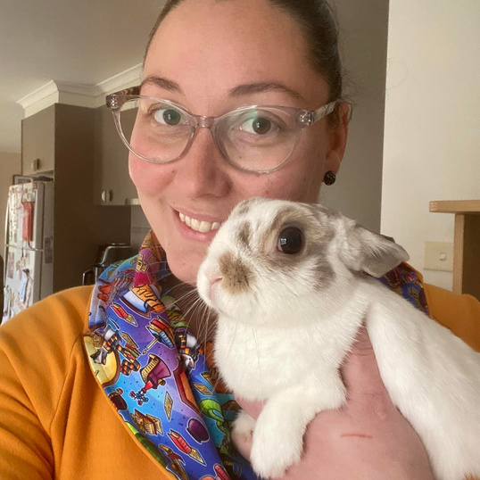 A white woman wearing glasses and a yellow jumper with a brightly coloured collar, cuddling a cute white rabbit with brown spots on it's face.