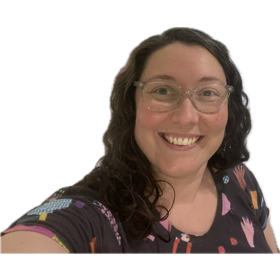 White lady with brown wavy hair, glasses, and a navy t-shirt with brightly coloured designs is smiling at the camera.